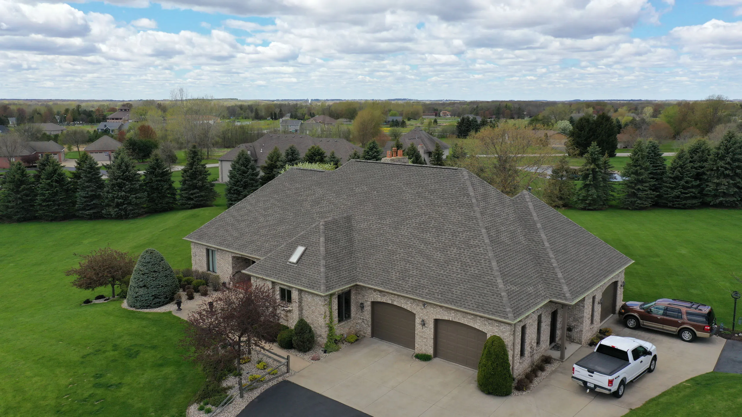 A large brick house with a multi-gabled gray roof, two-car garage, two parked vehicles, and a well-maintained green lawn with trees under a cloudy sky.